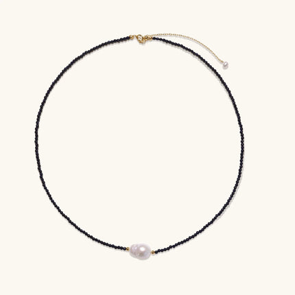 Delicate necklace with black blue sandstone beads and baroque pearl center on white background with gold accents.