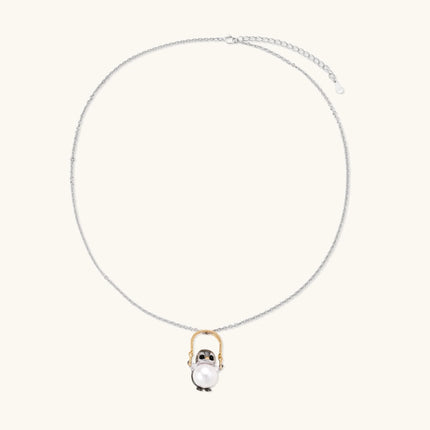 Whimsical owl pendant holding a white pearl on a gold swing, rhodium-plated silver chain on cream background.